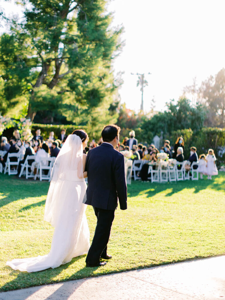 Bride in white dress and veil walks with a man in a suit down a sunlit garden path towards seated guests.