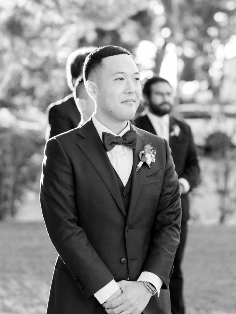 A groom in a suit with a bow tie stands outdoors, looking ahead calmly. Sunlight softly illuminates him, while blurred guests wait behind.