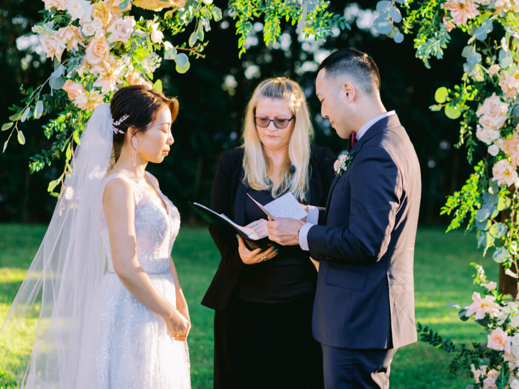 A bride and groom stand under a floral arch during a wedding ceremony. The officiant reads from a book. Sunlight filters through lush greenery.