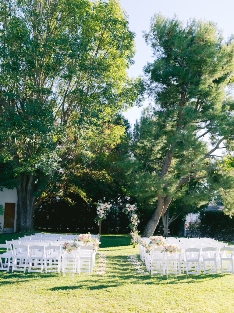 Outdoor wedding ceremony setup under tall trees, featuring an arch adorned with flowers. Rows of white chairs line the aisle.
