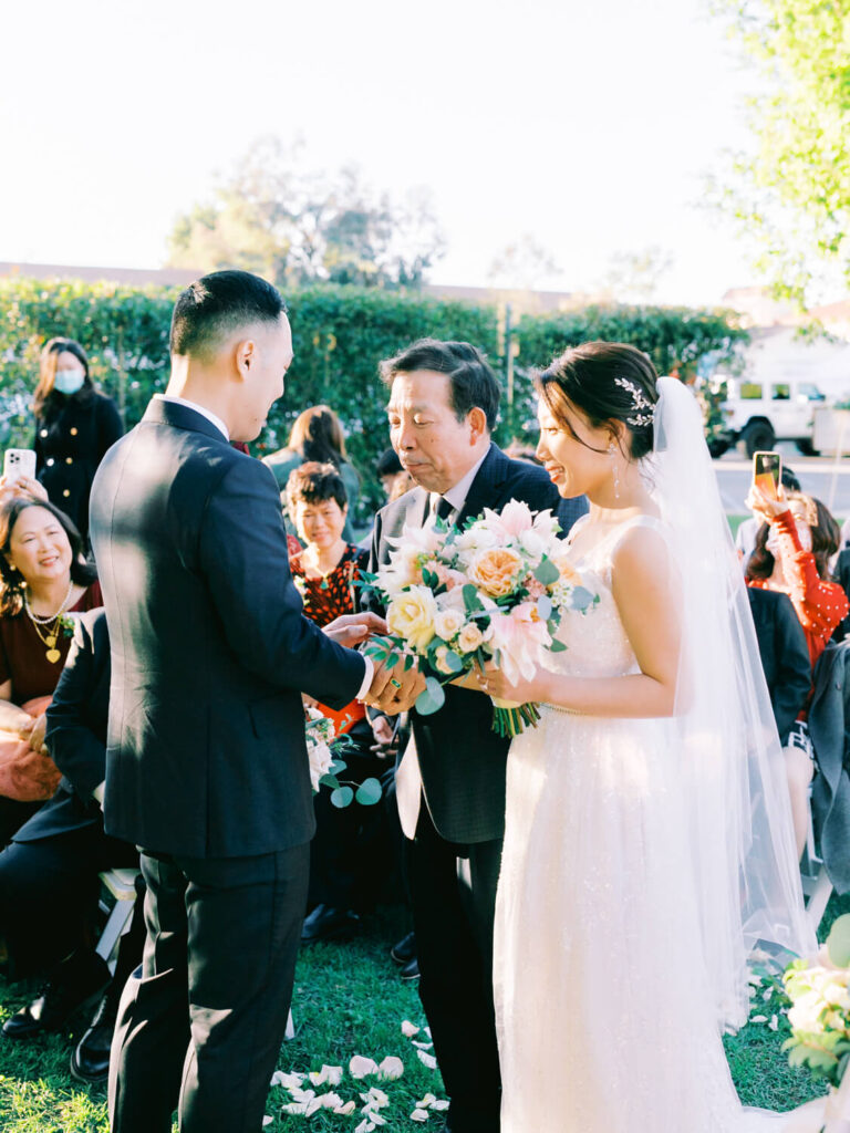 A bride in a white gown smiles as her father passes her hand to the groom during an outdoor wedding ceremony. Guests watch and capture moments with their phones.