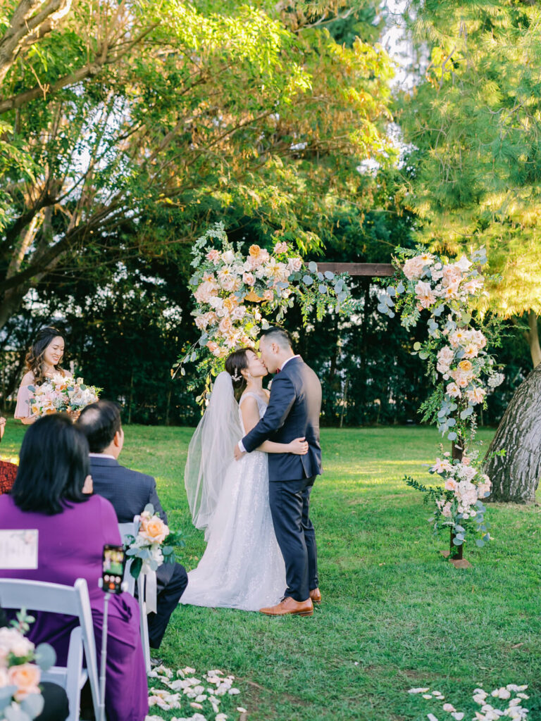 A couple kisses under a flower-adorned arch during an outdoor wedding. Guests seated nearby, lush green trees.