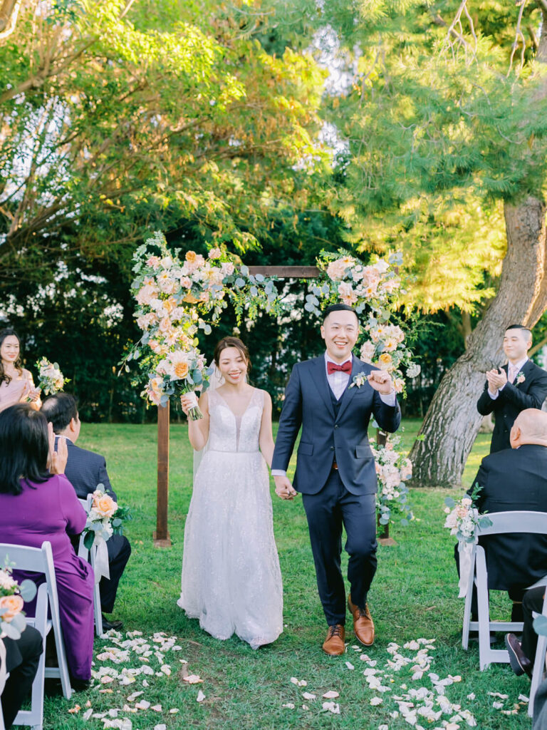 A joyful bride and groom walk hand in hand under a floral arch, surrounded by guests in an outdoor setting.