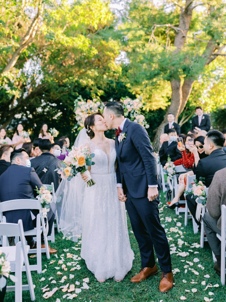 Bride and groom share a kiss in the aisle, surrounded by guests on white chairs. Blossoms and sunlight enhance the joyous outdoor wedding scene.