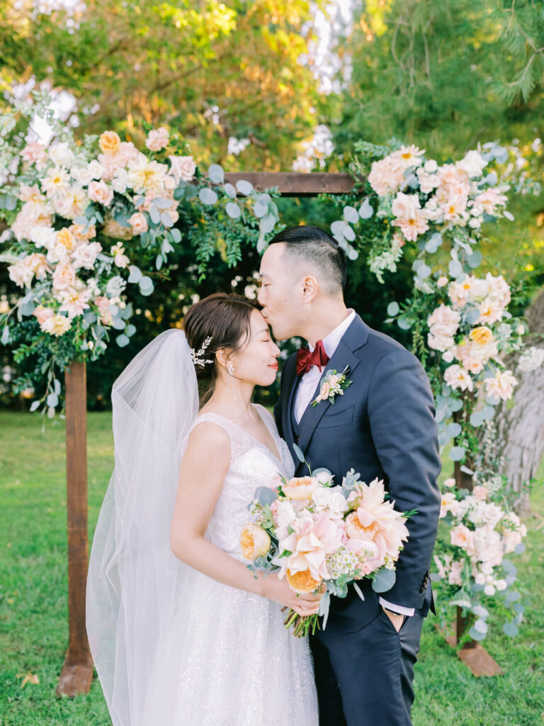 A bride in a white gown and veil holds a bouquet as the groom in a navy suit kisses her forehead. They're standing under a floral arch outdoors.