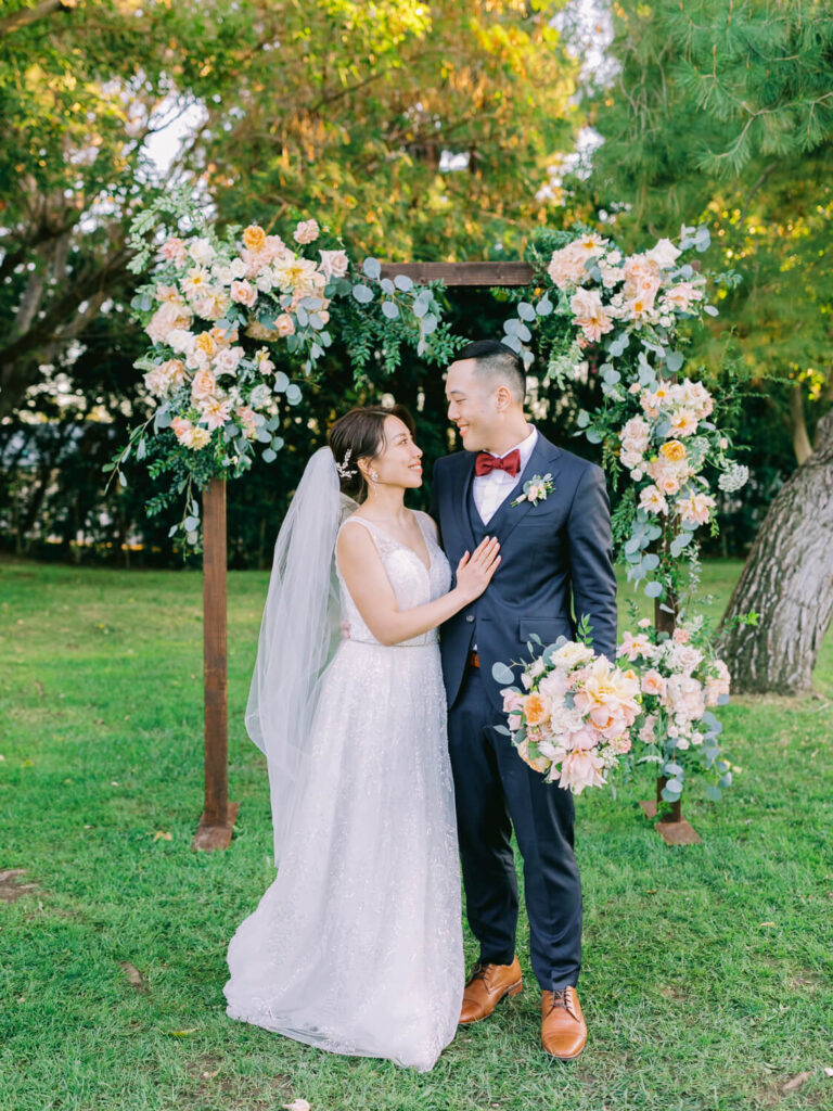 A bride in a white gown and veil and a groom in a dark suit with a red bow tie stand under a floral arch. They gaze lovingly at each other in a lush garden setting.