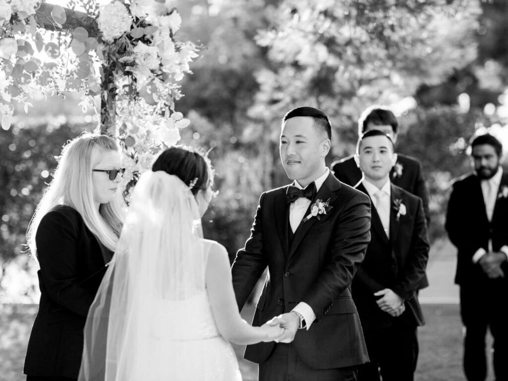 A joyful wedding scene shows a couple holding hands under a floral arch. The groom smiles warmly at the bride while a person officiates the ceremony. Black and white photo.