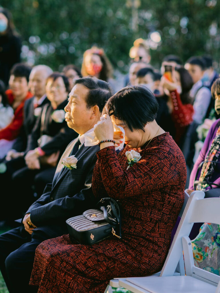 A woman in a red dress wipes tears with a tissue at an outdoor event. She's sitting next to a man in a suit.