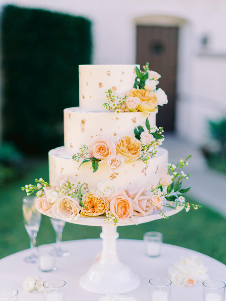 Three-tiered wedding cake adorned with soft peach and white roses, green foliage, and gold leaf accents, on a white stand.