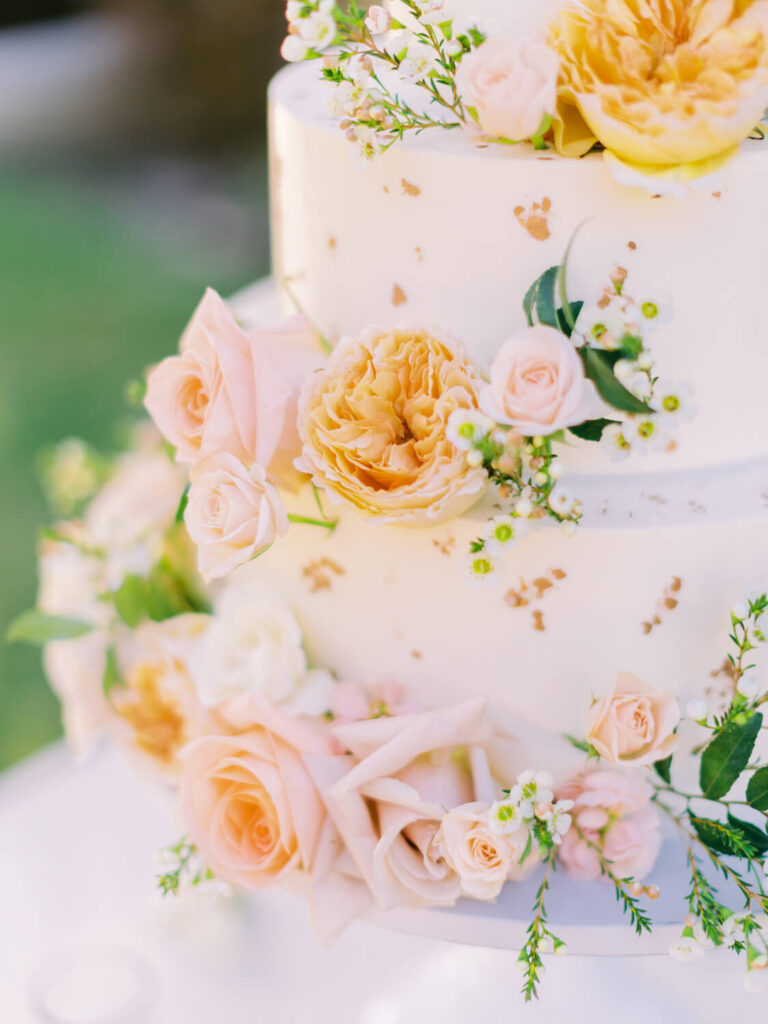 Three-tier white cake adorned with peach, pink, and white roses, green leaves, and delicate white blossoms.