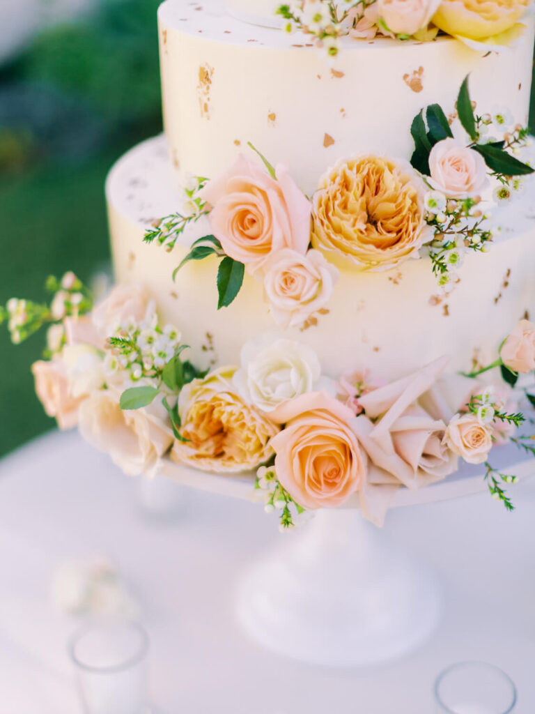 Three-tier white cake adorned with peach, pink, and white roses, green leaves, and delicate white blossoms.
