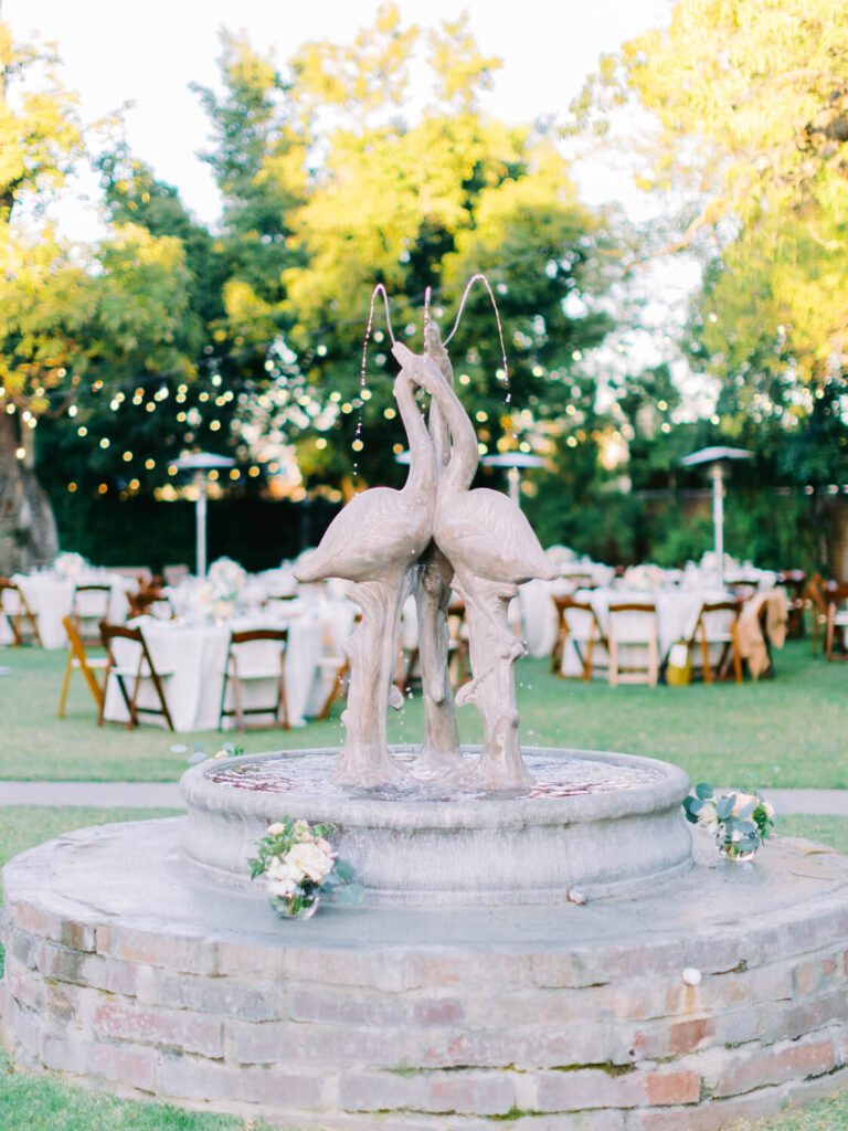 Stone fountain with bird sculptures, spouting water, set in a garden with round tables and chairs. Fairy lights hang above.