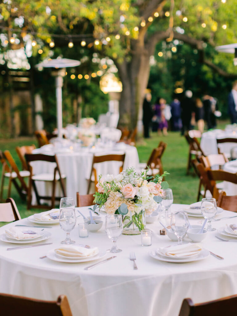 Outdoor wedding reception with round tables, elegantly set with white linens and floral centerpieces. String lights twinkle above in a serene garden setting.