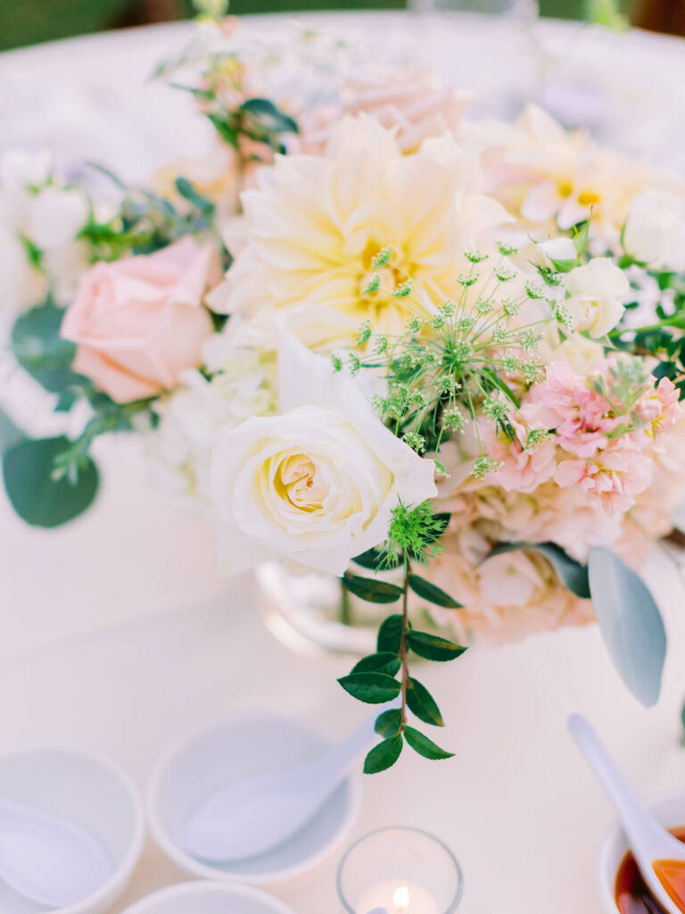 A delicate floral arrangement with pale pink and white roses, greenery, and soft yellow blossoms in a glass vase, set on a light table with small bowls and a candle.