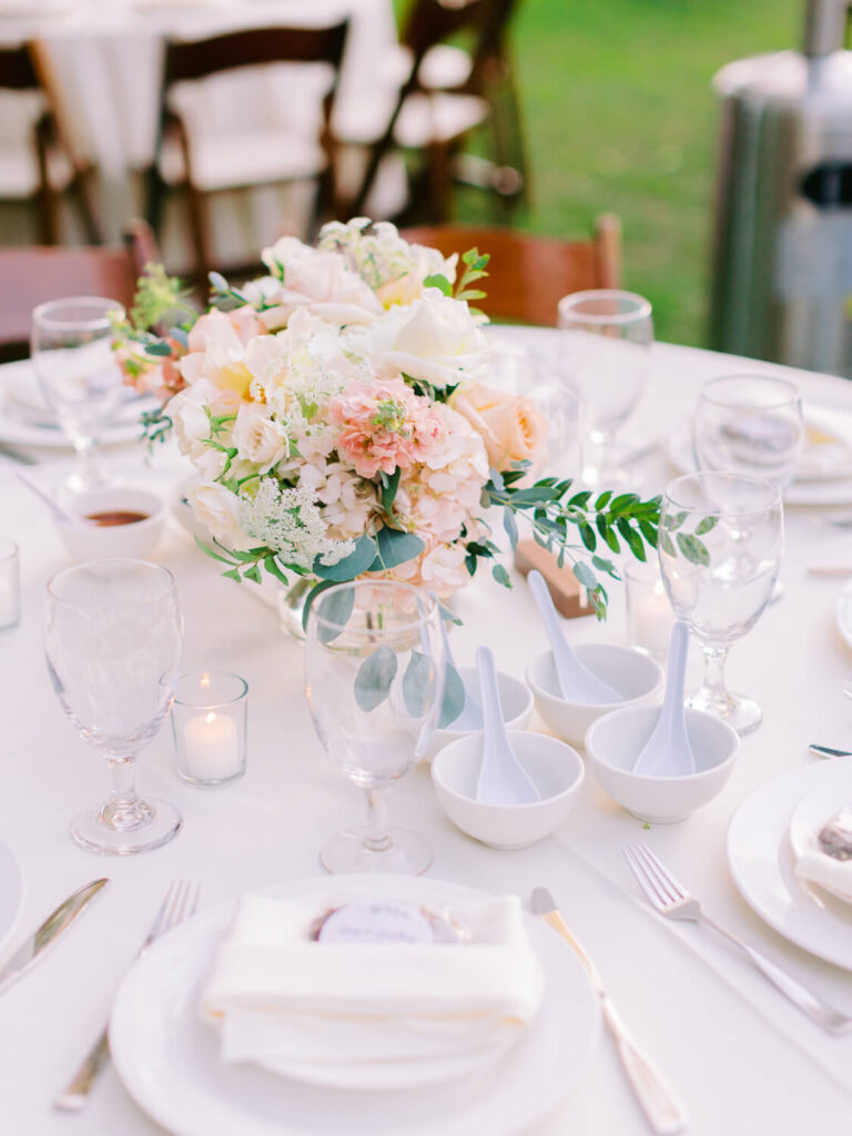 Elegant wedding table setting with a centerpiece of pastel flowers, surrounded by glassware, white bowls with spoons, and flickering candles, creating a romantic ambiance.