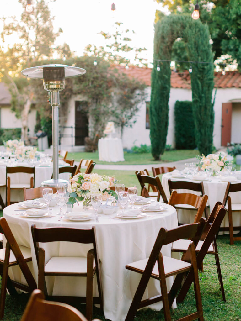 Outdoor wedding reception with round tables covered in white cloths, wooden chairs, floral centerpieces, and a patio heater.