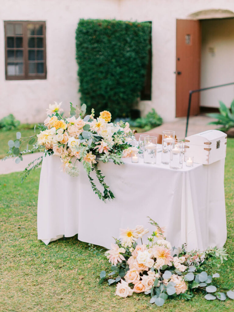 Elegant outdoor wedding setup with a white table adorned with peach and white floral arrangements. Glass candles add a romantic touch against a garden backdrop.
