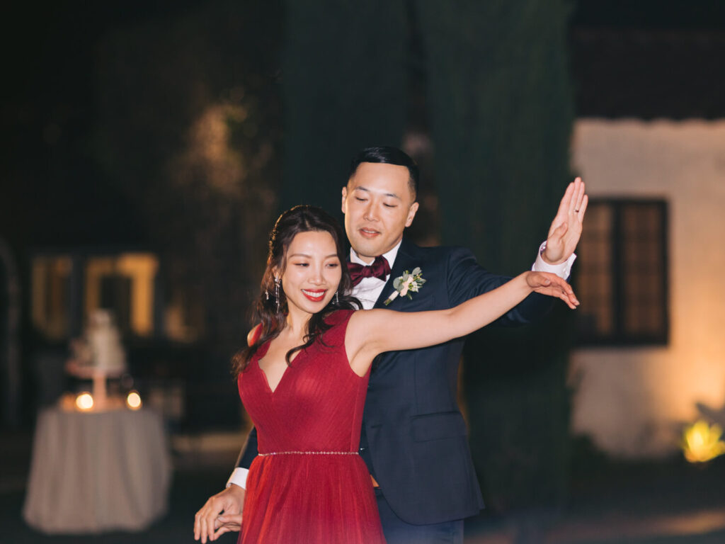 A couple gracefully dances outdoors at night. The woman in a red dress smiles, while the man in a suit guides her.