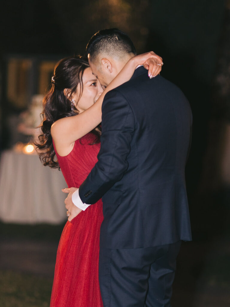 A couple dances closely, the woman in a red dress, and the man in a dark suit. Their embrace is intimate and emotional, set against a blurred, dimly-lit backdrop.