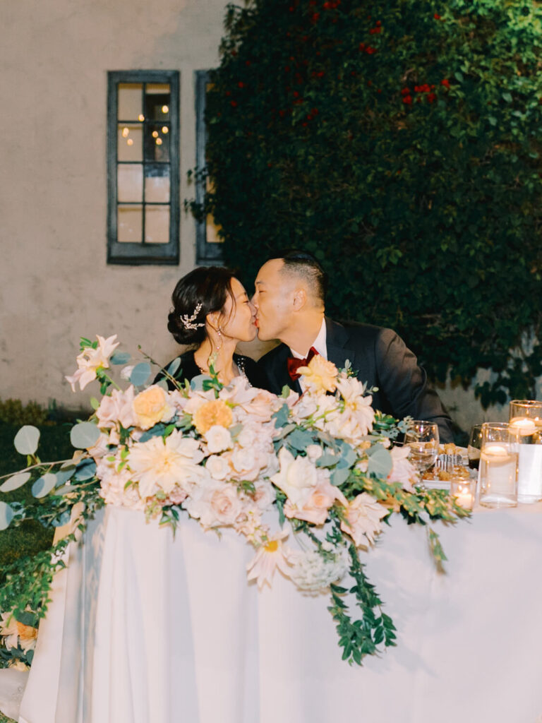 A couple kisses behind a table adorned with lush pastel flowers and candles. The scene is romantic with a backdrop of ivy-covered walls and soft lighting.