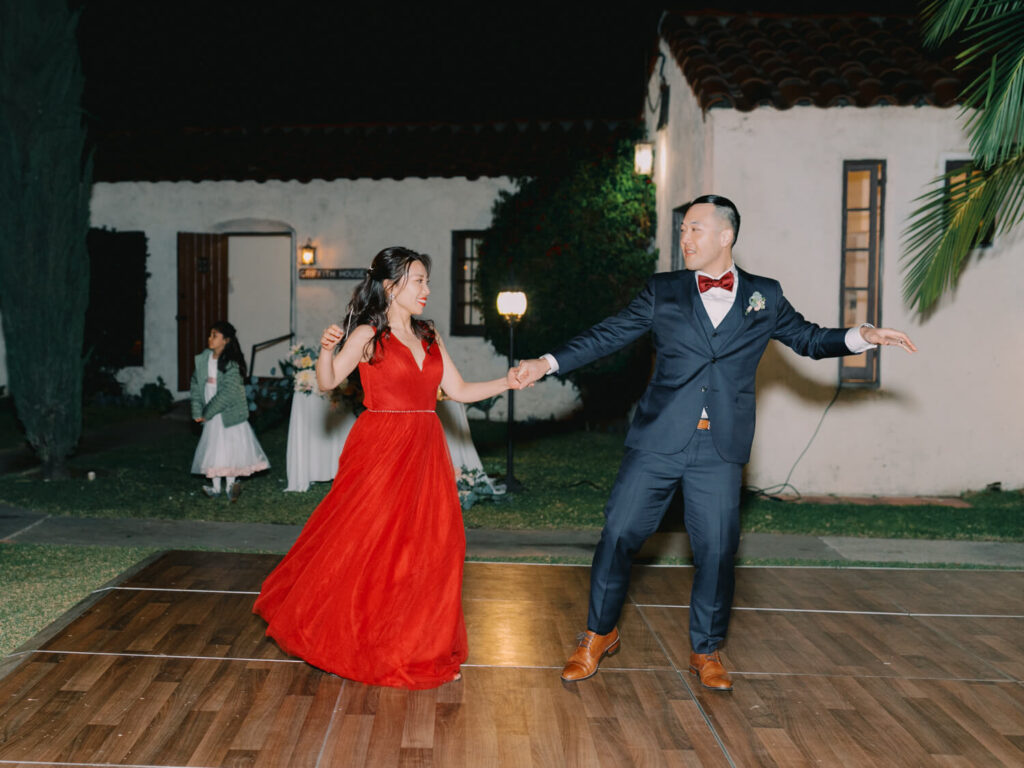 A couple dances joyfully on an outdoor wooden floor at night. The woman wears a flowing red dress, and the man is in a navy suit.