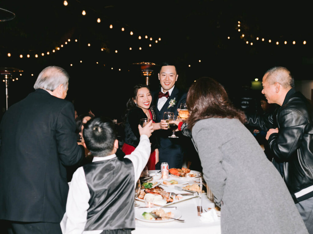 A joyful group celebrates at an outdoor event under string lights. People toast with drinks around a table filled with food.