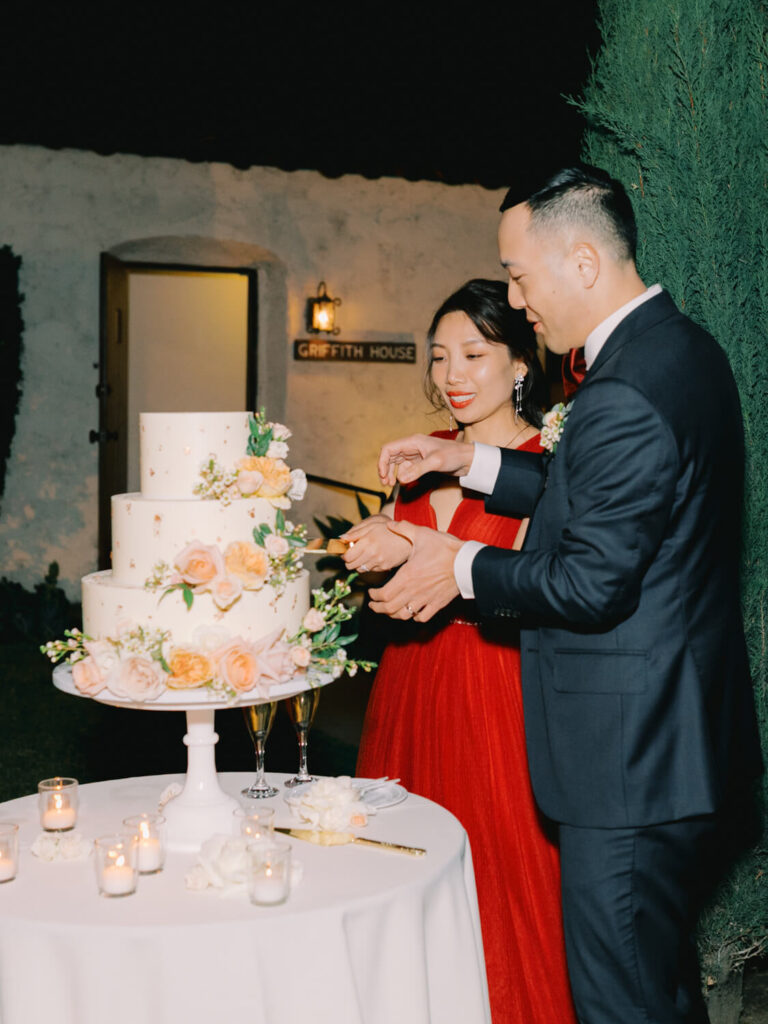 A couple in elegant attire cuts a three-tiered wedding cake adorned with peach flowers.