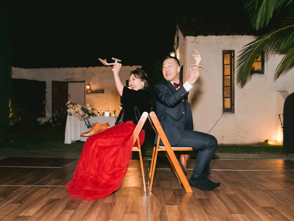 A couple sits back-to-back on chairs at a wedding reception, each holding up a shoe and laughing.
