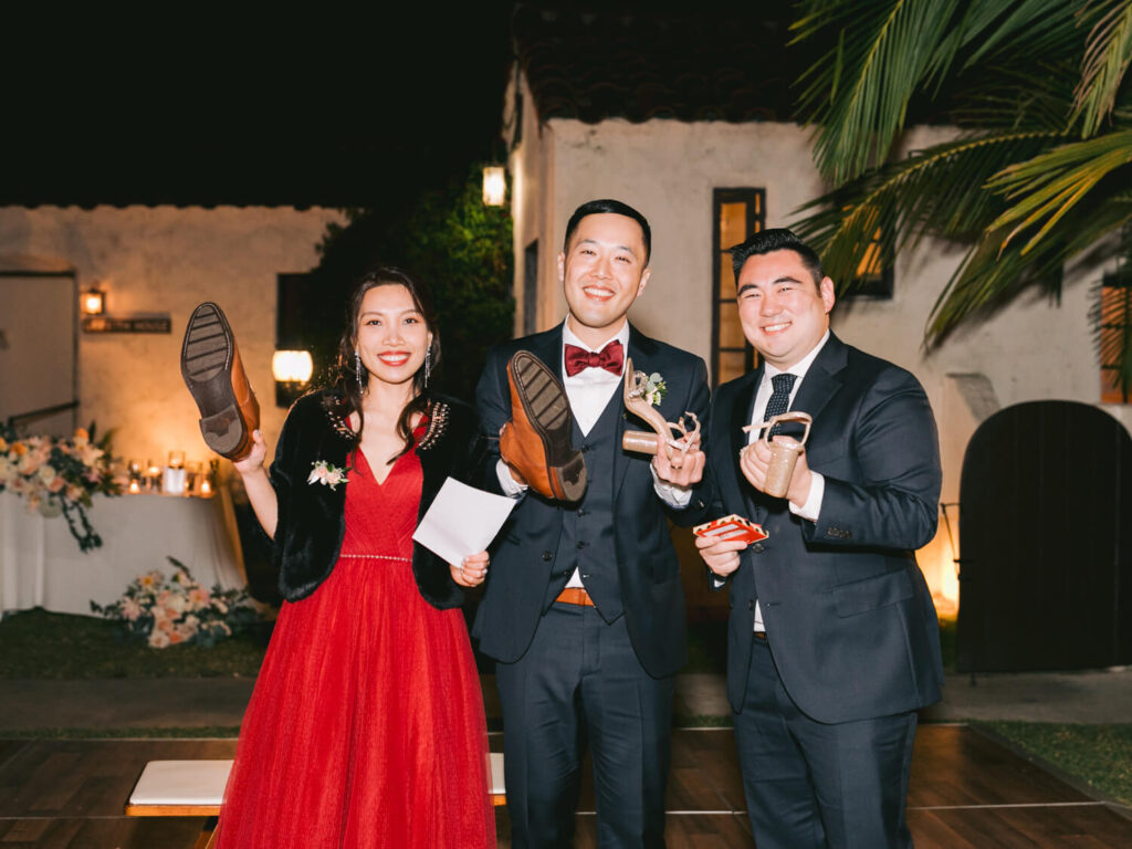 A joyful group of three people poses at an outdoor evening event. Two hold shoes, and one holds cards.