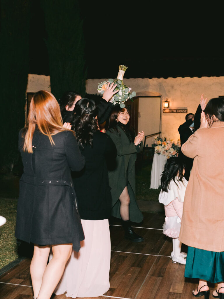 A group of women and a child celebrate outdoors at night. One woman catches a bouquet, surrounded by clapping guests.