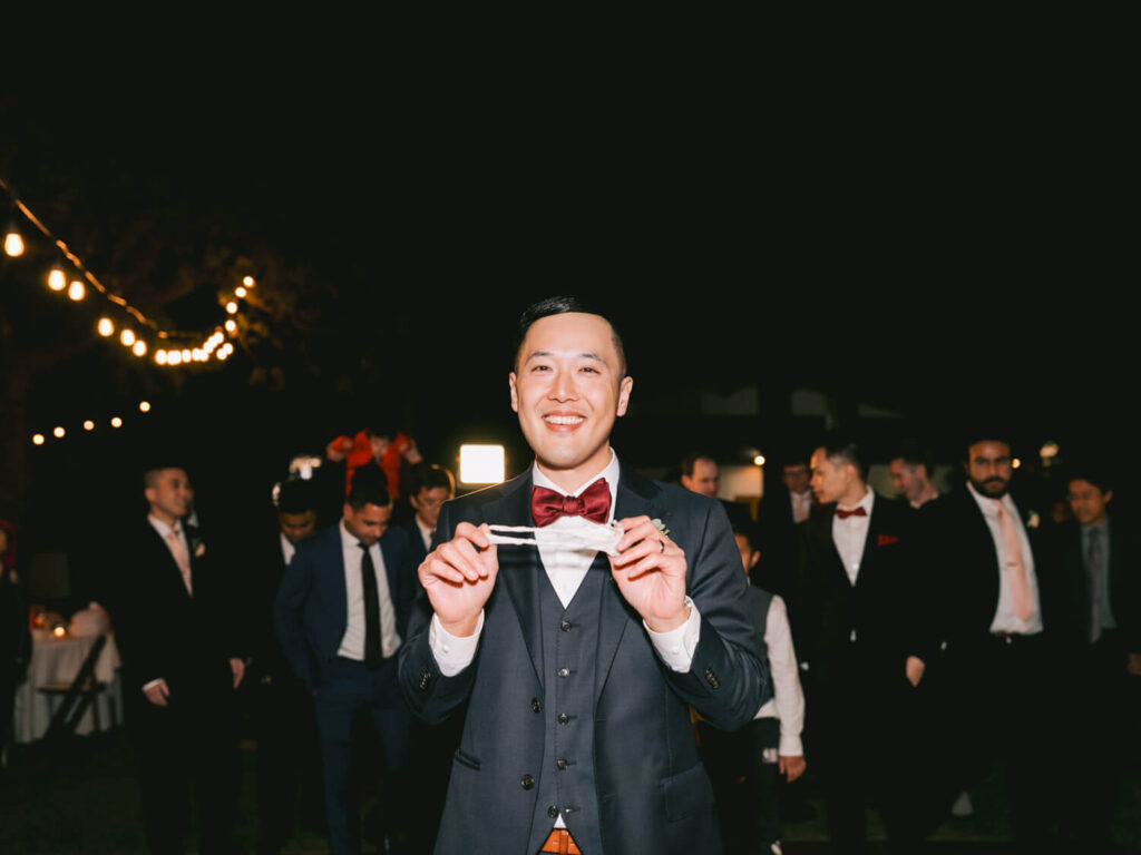 A man in a suit and bow tie stands smiling, holding a garter. He is surrounded by men in suits under string lights at a nighttime reception.
