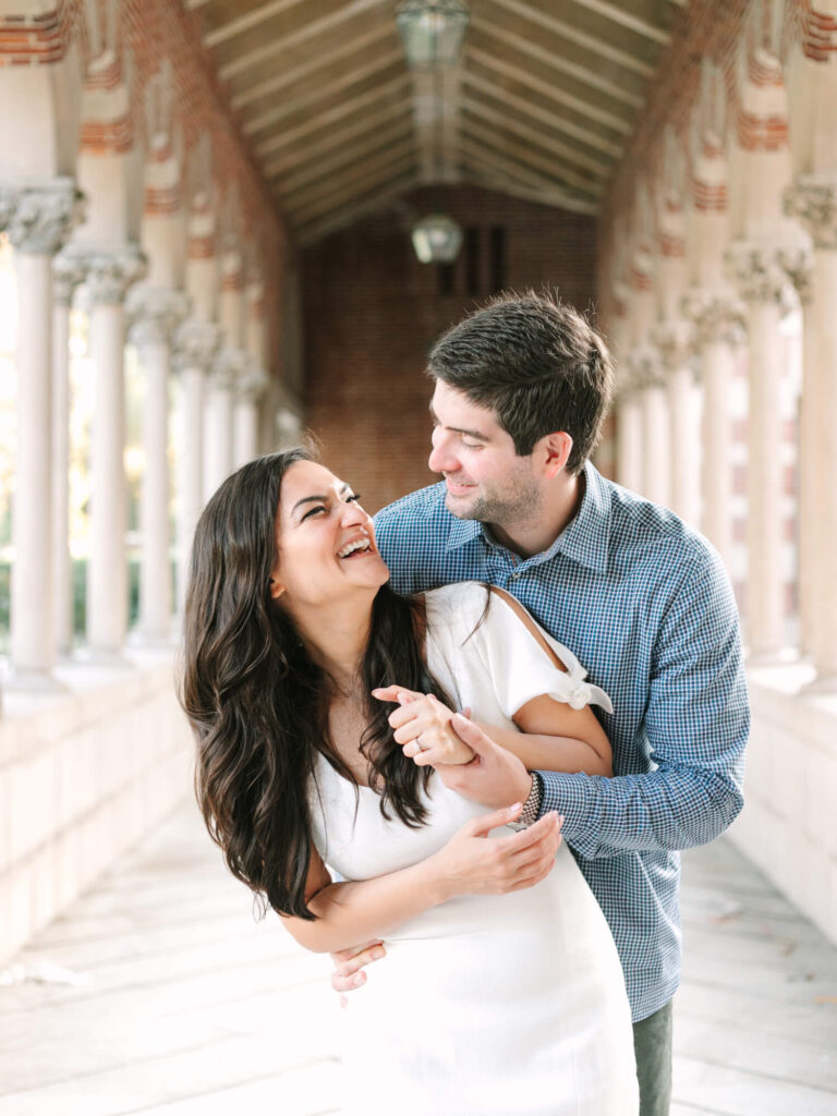 A joyful couple laughs together in a stone corridor with ornate columns. The woman wears a white dress, the man a blue checkered shirt.