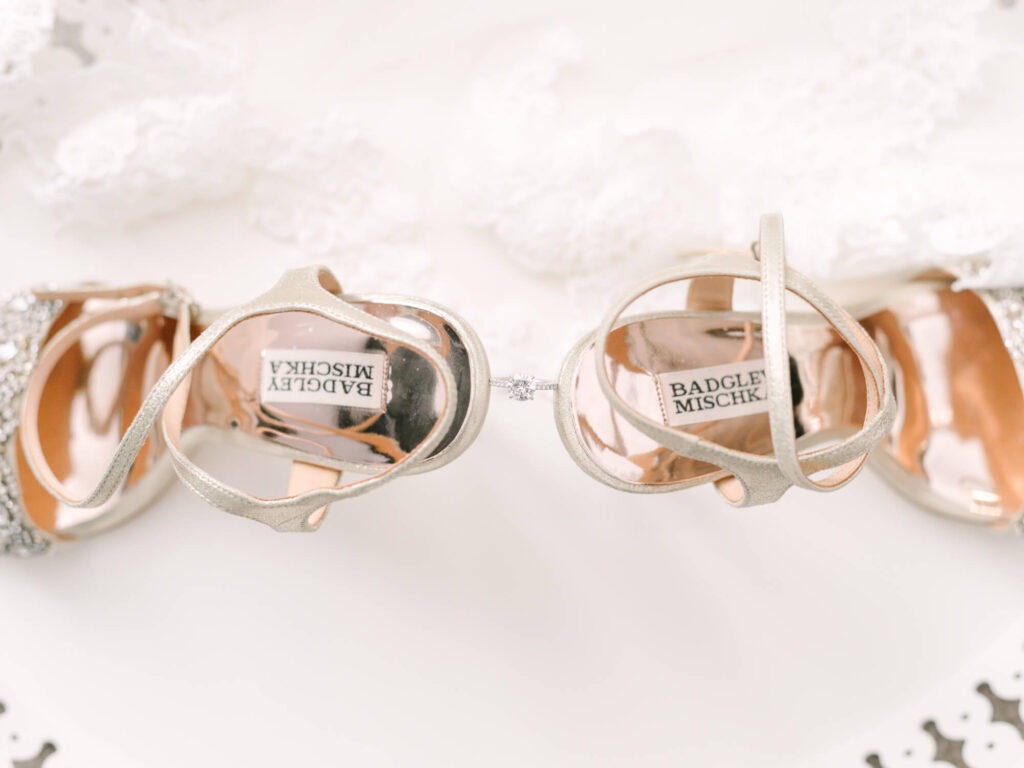 Elegant silver Badgley Mischka high heels with intricate detailing, displayed overhead on a light lace fabric.