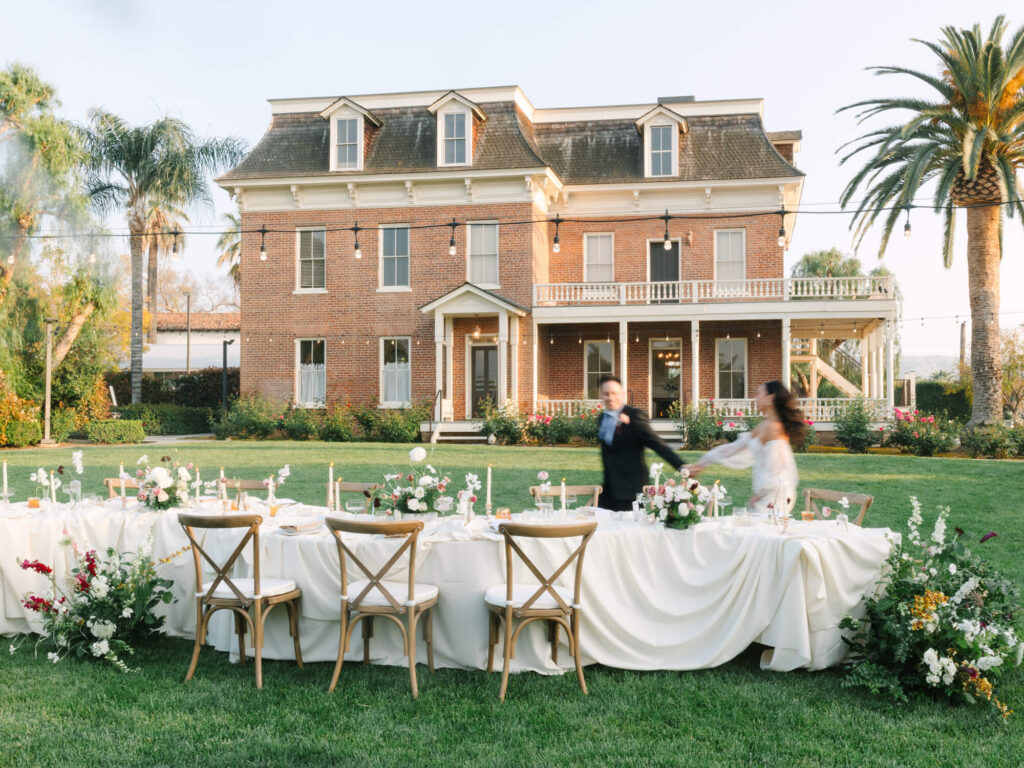 A beautifully set outdoor table in front of a large, elegant brick house with a manicured garden. A couple joyfully moves past.