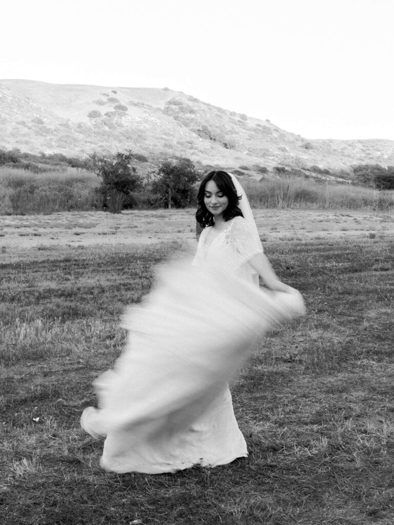 A black and white photo of a bride joyfully twirling outdoors. Her flowing gown creates a sense of movement against a backdrop of rolling hills and grass.