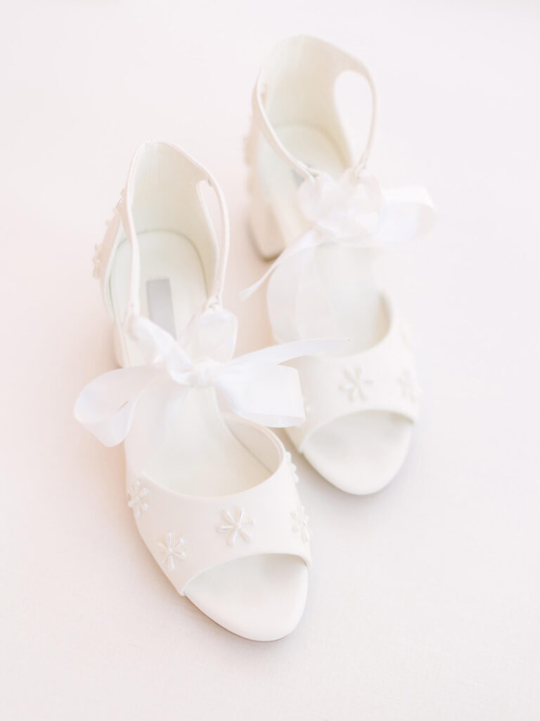 Elegant white bridal shoes with open toes and ankle straps, adorned with small floral patterns and sheer ribbon bows, resting on a soft white surface.