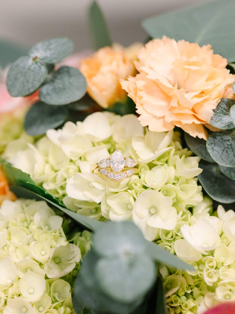 A sparkling diamond ring nestled among light green hydrangeas with soft peach carnations and eucalyptus leaves.