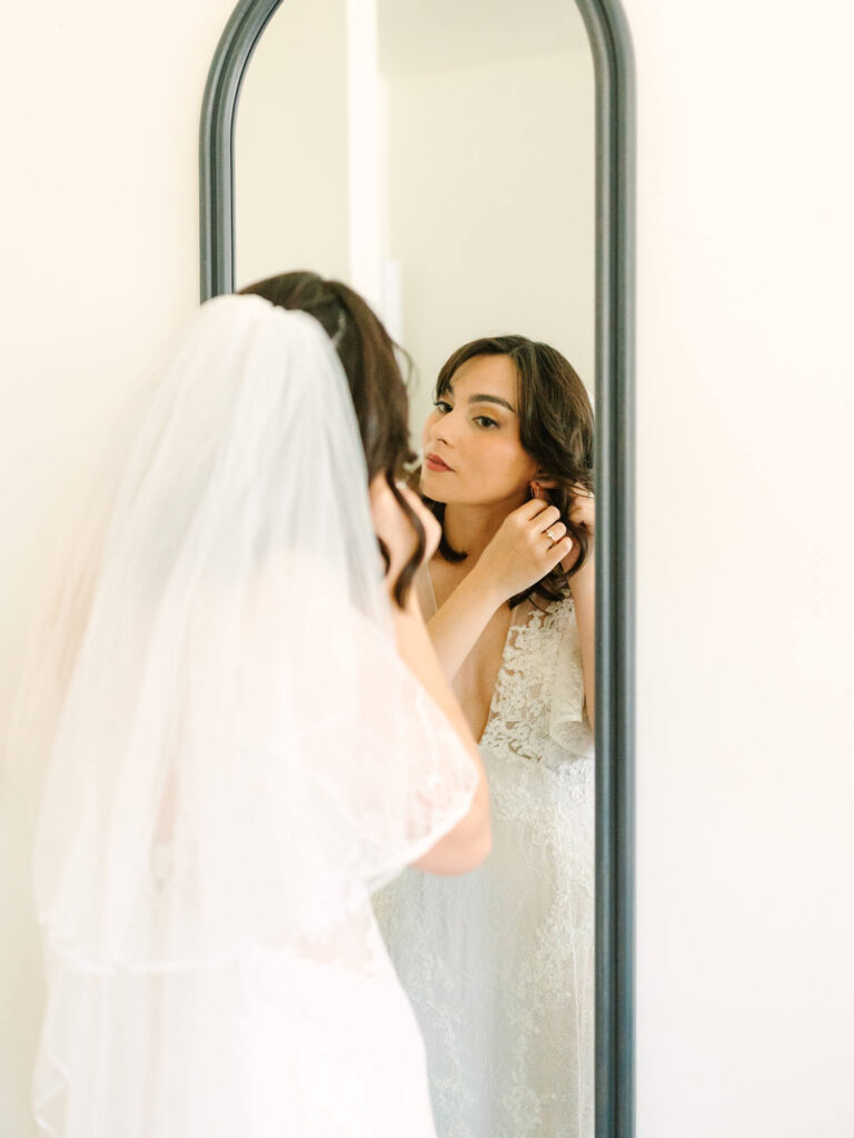 A bride in a white lace dress and veil adjusts her earrings, gazing into a mirror.