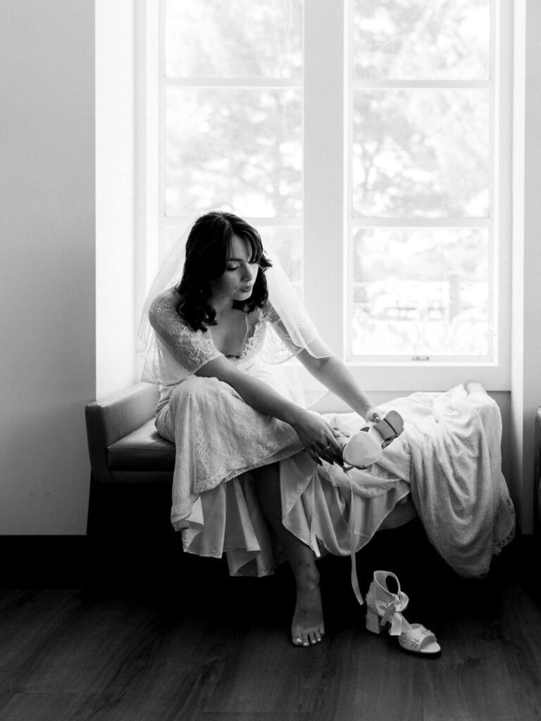 A bride in a lace dress and veil sits by a large window, adjusting her shoe.