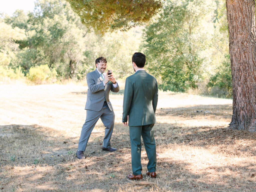 Two men in suits outdoors; one man energetically takes a photo of the other, who stands with his back to the camera, under a tree in a sunlit field.