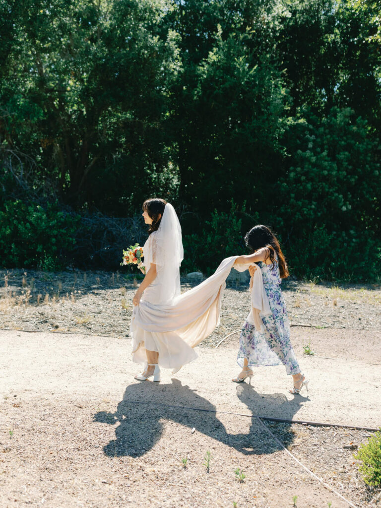 Bride walking outdoors in sunlight, holding a bouquet, while a young girl assists with her train.