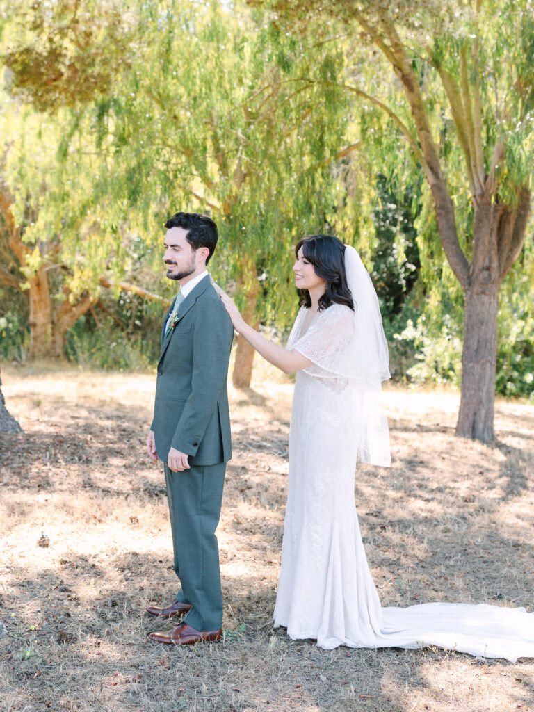 A bride in a white gown gently touches a groom's shoulder from behind in a sunlit, leafy park.