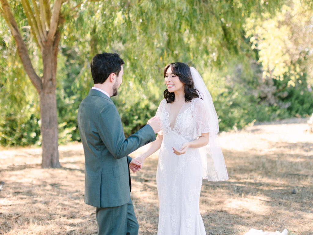 A bride in a lace gown and veil smiles warmly at a groom in a green suit under a tree.