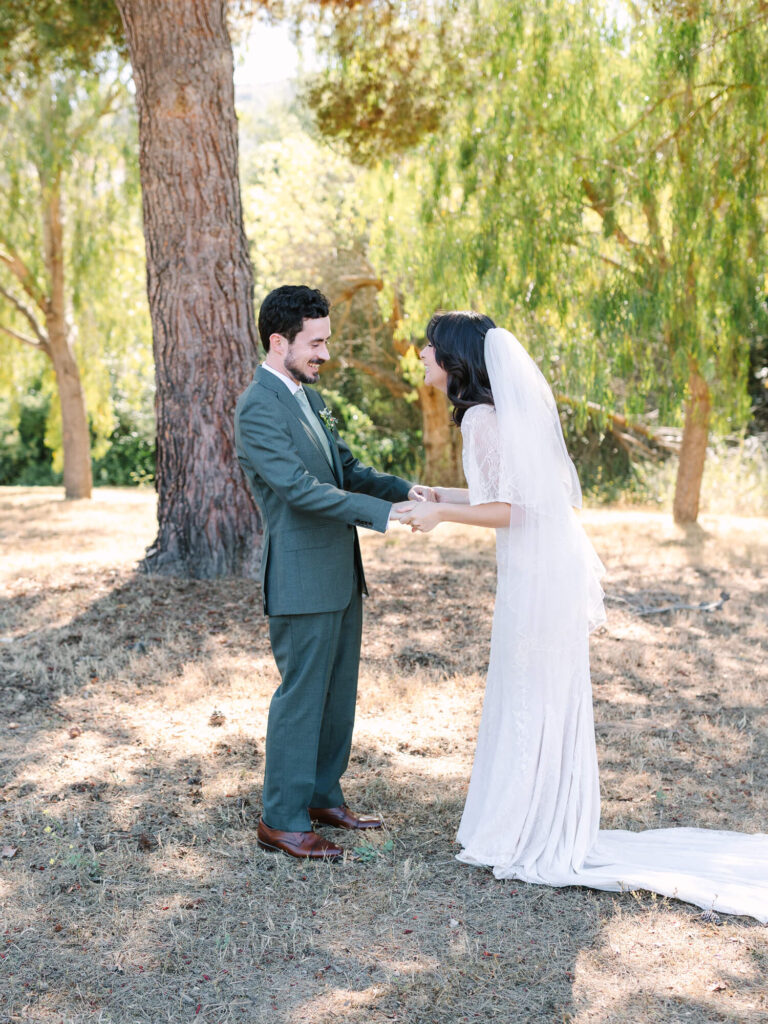 Bride in a lace gown and veil holds hands with a groom in a green suit in a sunlit, wooded park.