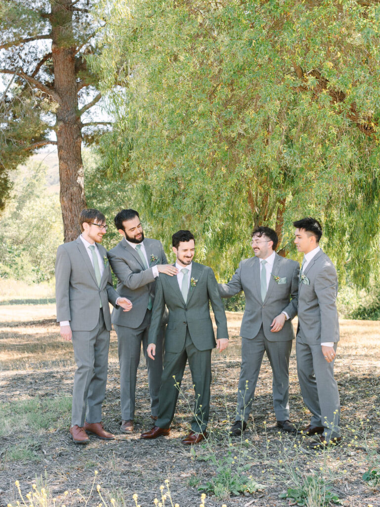 Five men in gray suits laugh and interact warmly outdoors, standing on grass under tall trees.