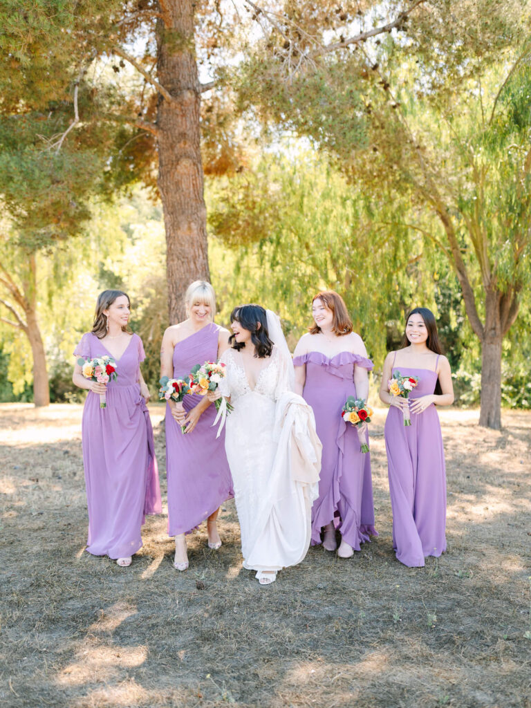 A bride in a white dress walks with four bridesmaids in lavender dresses, holding bouquets, under a canopy of trees.