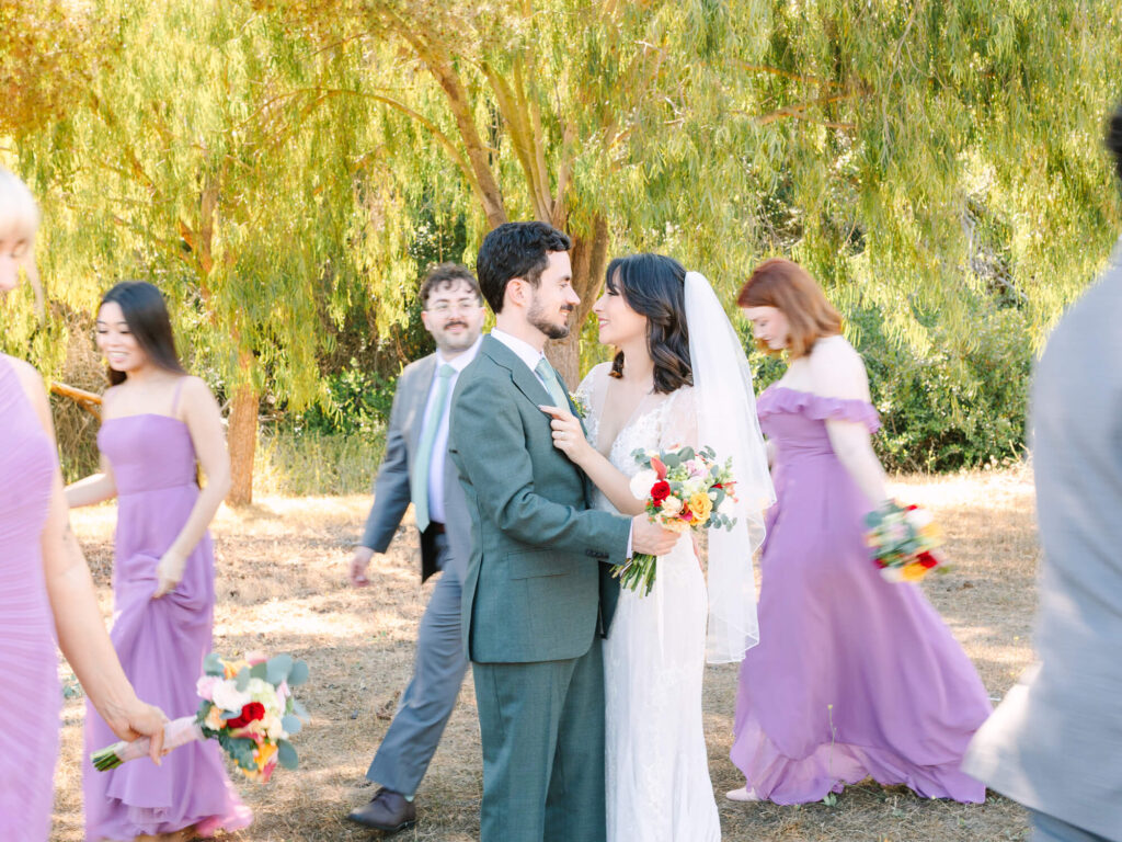 Bride and groom smile at each other under sunny trees, surrounded by bridesmaids in lavender dresses.