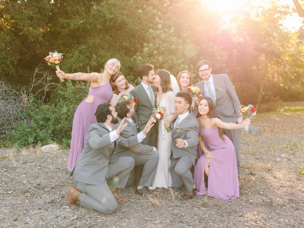 A joyful wedding party in a sunlit forest setting. Bride and groom kiss at the center, surrounded by smiling bridesmaids in lilac and groomsmen in gray suits, holding bouquets.