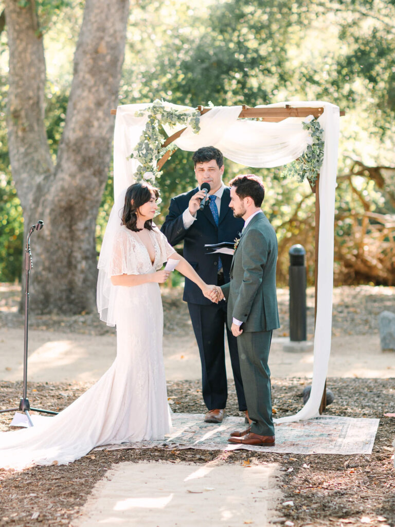 Bride in white gown and groom in grey suit hold hands under a wooden arch adorned with greenery. An officiant, in a suit, speaks into a microphone.