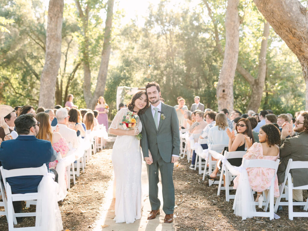 Bride in white dress and groom in gray suit stand smiling, holding hands under dappled sunlight. Guests seated on white chairs applaud in a wooded outdoor setting.